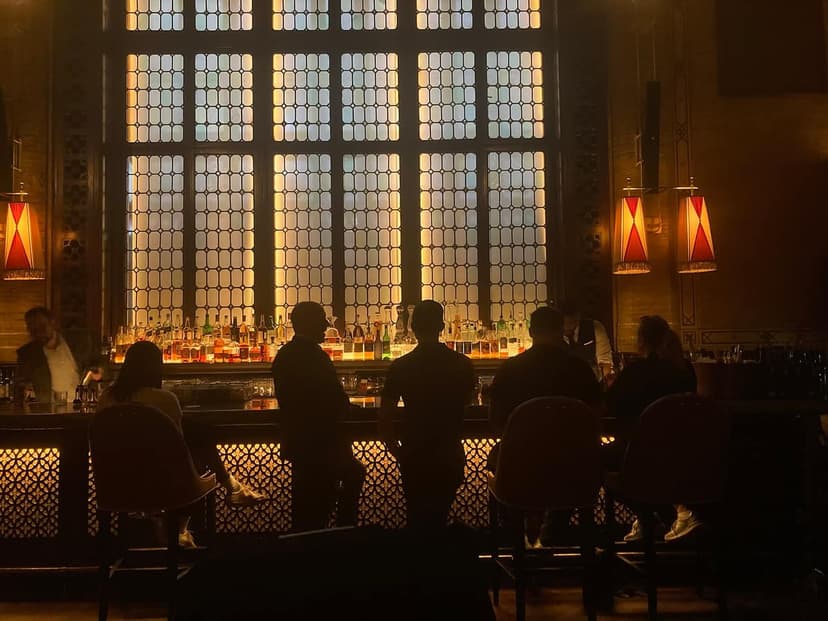 Interior view of a bar with a large window featuring many small panes, a long bar counter with bottles, and several patrons seated or standing at