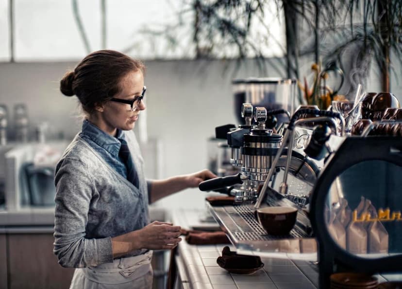 A barista with glasses and a bun hairstyle operates an espresso machine in a cafe. A coffee cup sits on the machine's drip tray.