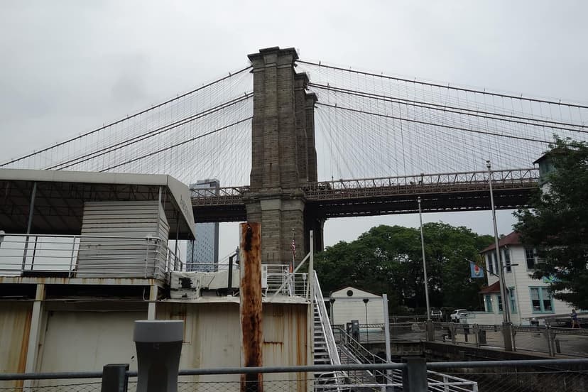 View of the Brooklyn Bridge from a park, showing its stone towers and suspension cables against a cloudy sky.