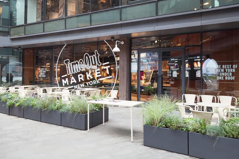 Exterior view of Time Out Market New York with its prominent sign, outdoor dining tables, chairs, and plant-filled planters.