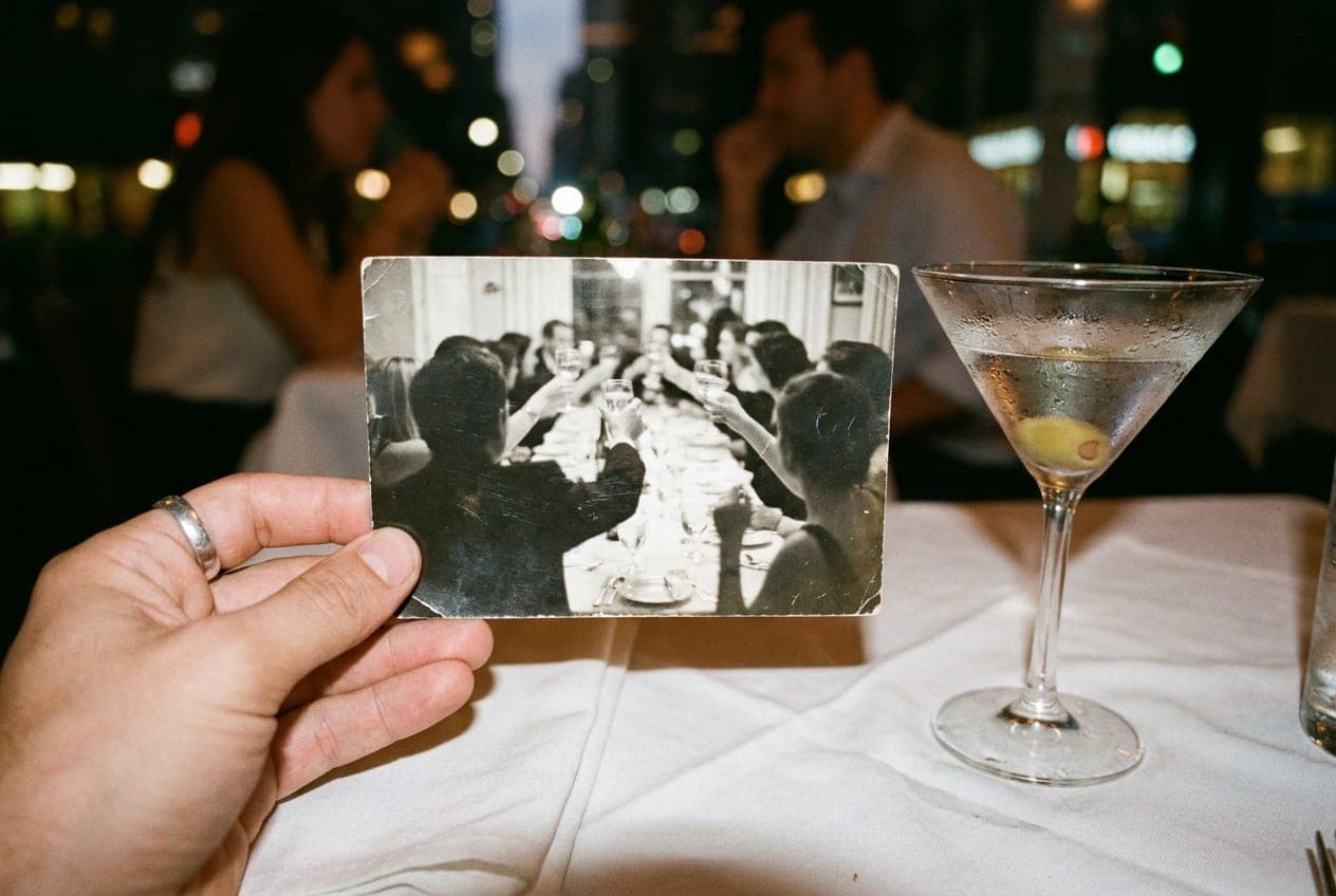 A hand holding a vintage photograph of a dinner party over a white tablecloth with a martini glass.