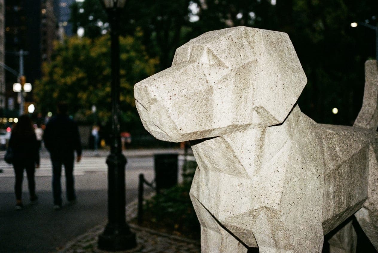 A large abstract dog sculpture made of concrete and steel standing in front of the Central Park entrance at night.