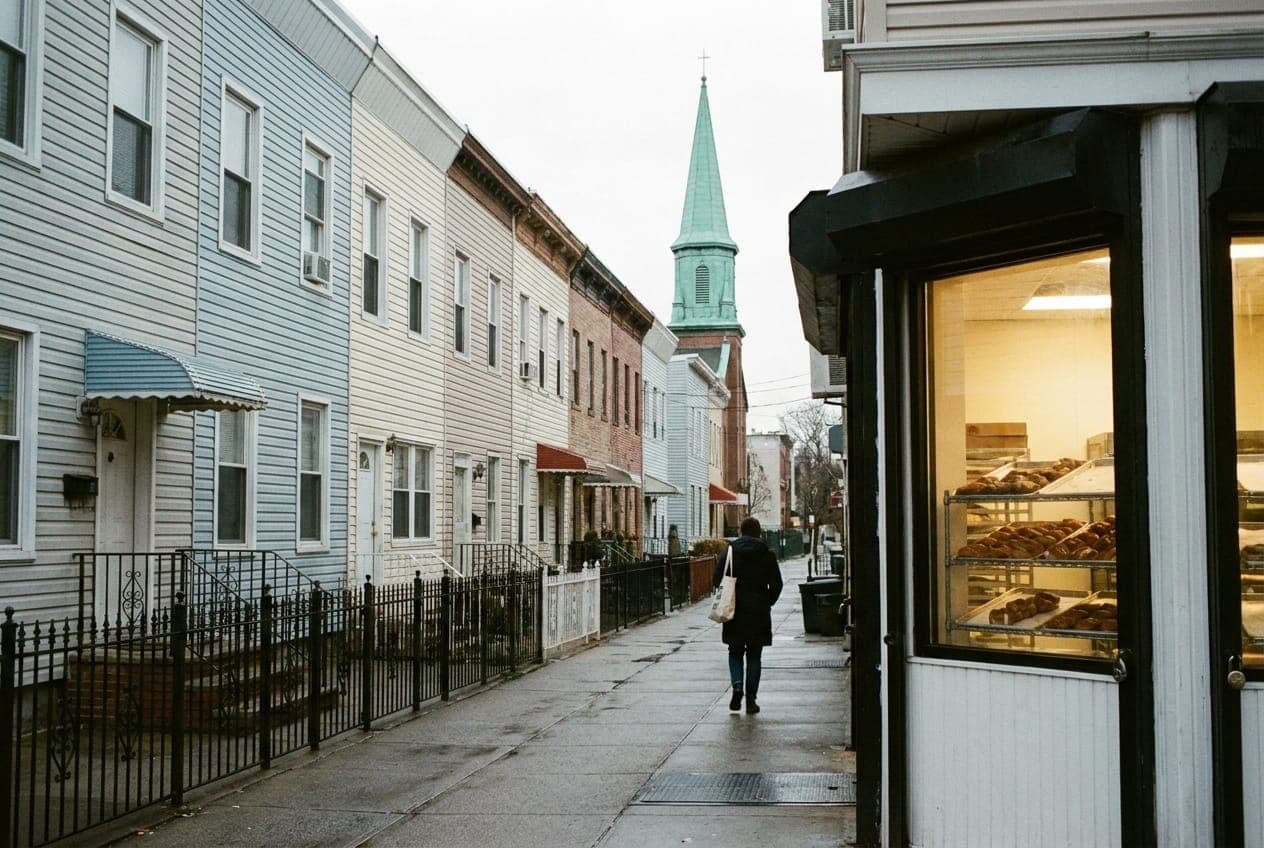 A quiet Greenpoint side street in the morning with vinyl-sided row houses, a church steeple and a bakery window glowing at the corner