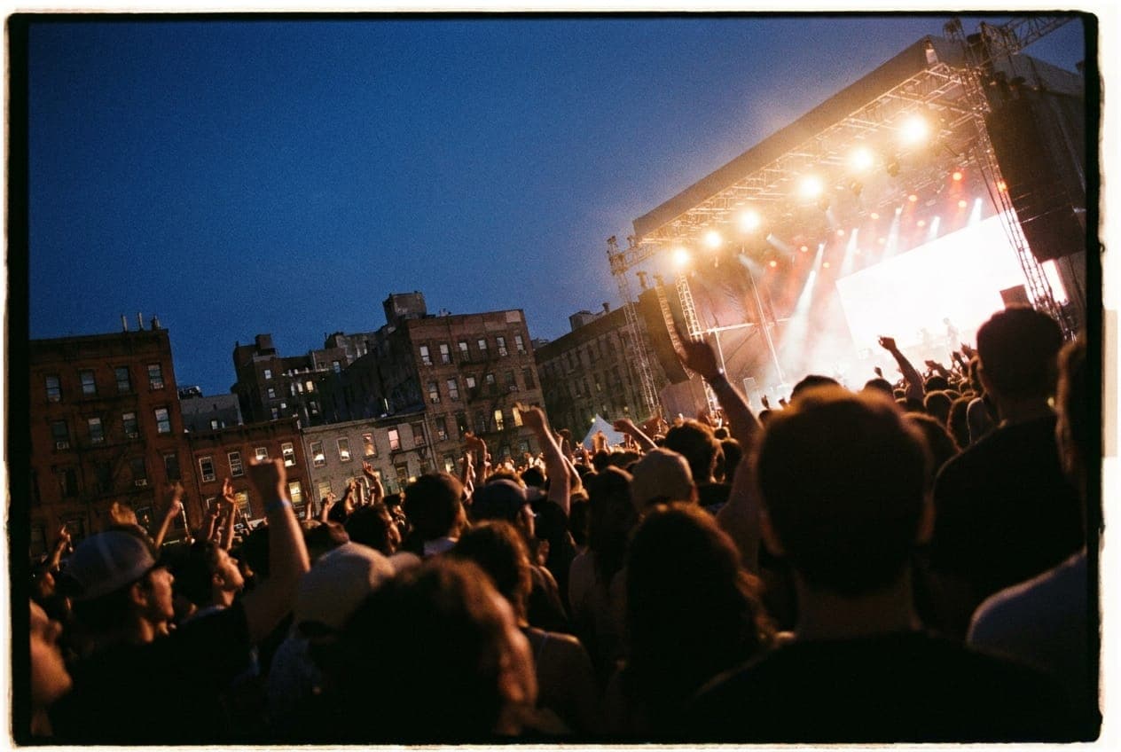 A crowd of people silhouetted against the bright lights of an outdoor concert stage at dusk in New York City.