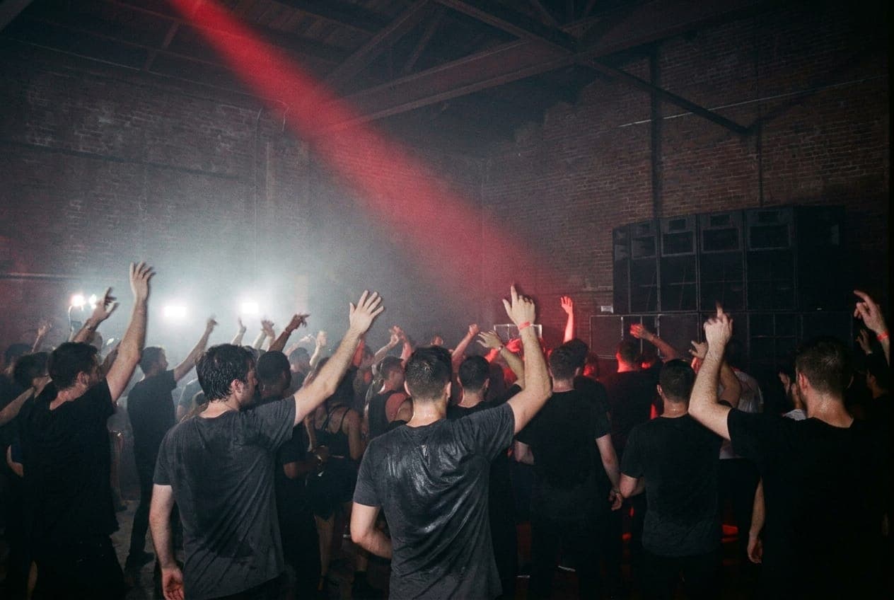 Silhouettes of dancers in a dark warehouse cut by a single red strobe beam and haze, arms raised, no faces visible